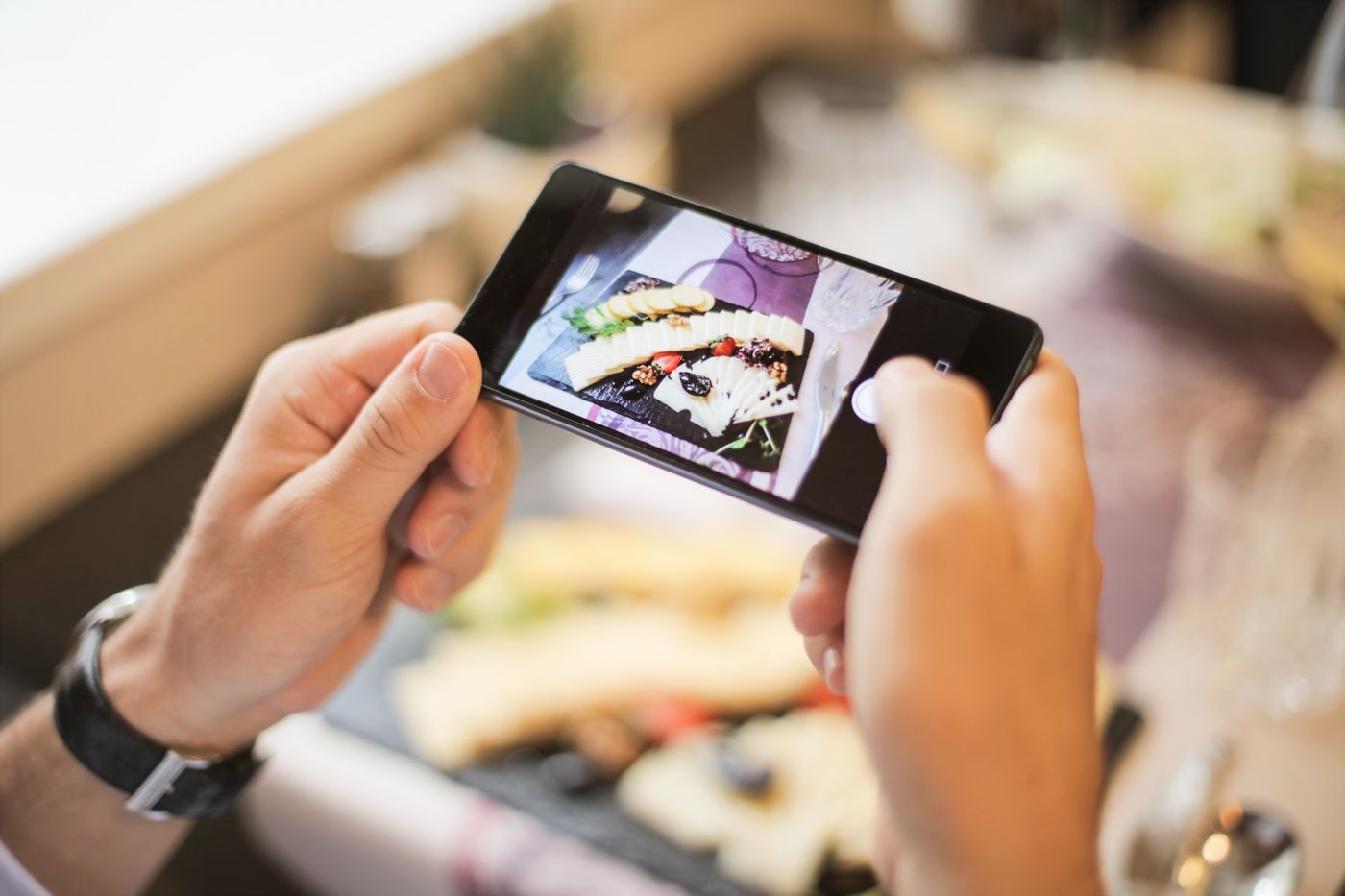 Propriétaire d'un petit commerce photographiant des plats dressés au smartphone près d'une fenêtre lumineuse, en suivant une liste de prises de vue simple
