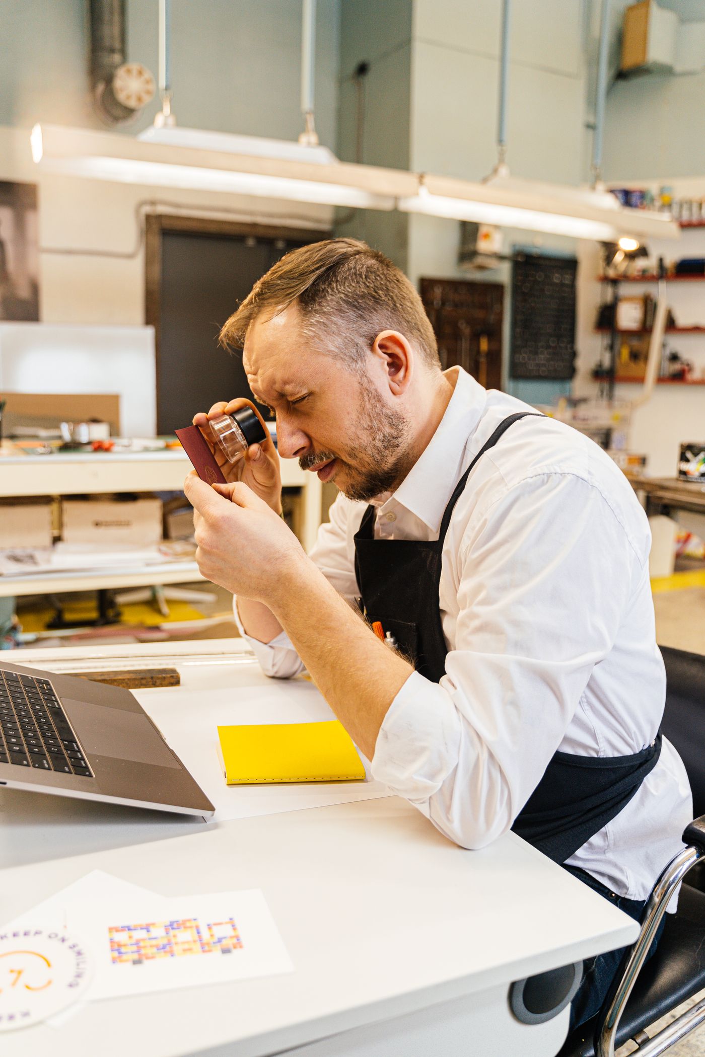 A man in a print studio holding a magnifier to his eye to inspect a printed sample closely at his desk