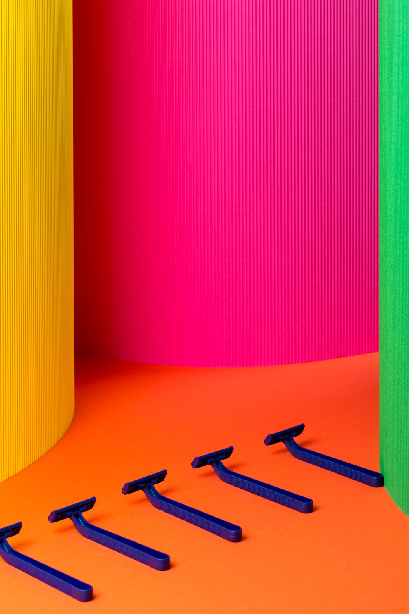 Five identical dark blue disposable razors laid out in an evenly spaced row on a flat orange surface in front of yellow, pink and green coloured panels