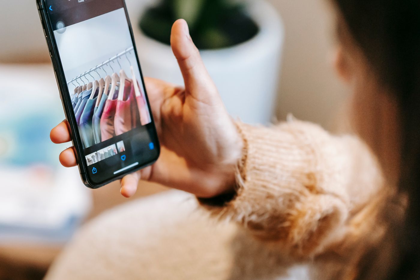 Over-the-shoulder close-up of a woman holding a smartphone showing a shopping app, with a photo of a clothing rail filling the screen and a strip of small thumbnails beneath it