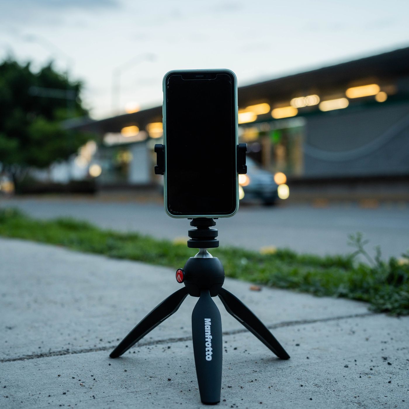 A smartphone clamped to a small tripod standing on a concrete pavement, aimed at a street scene
