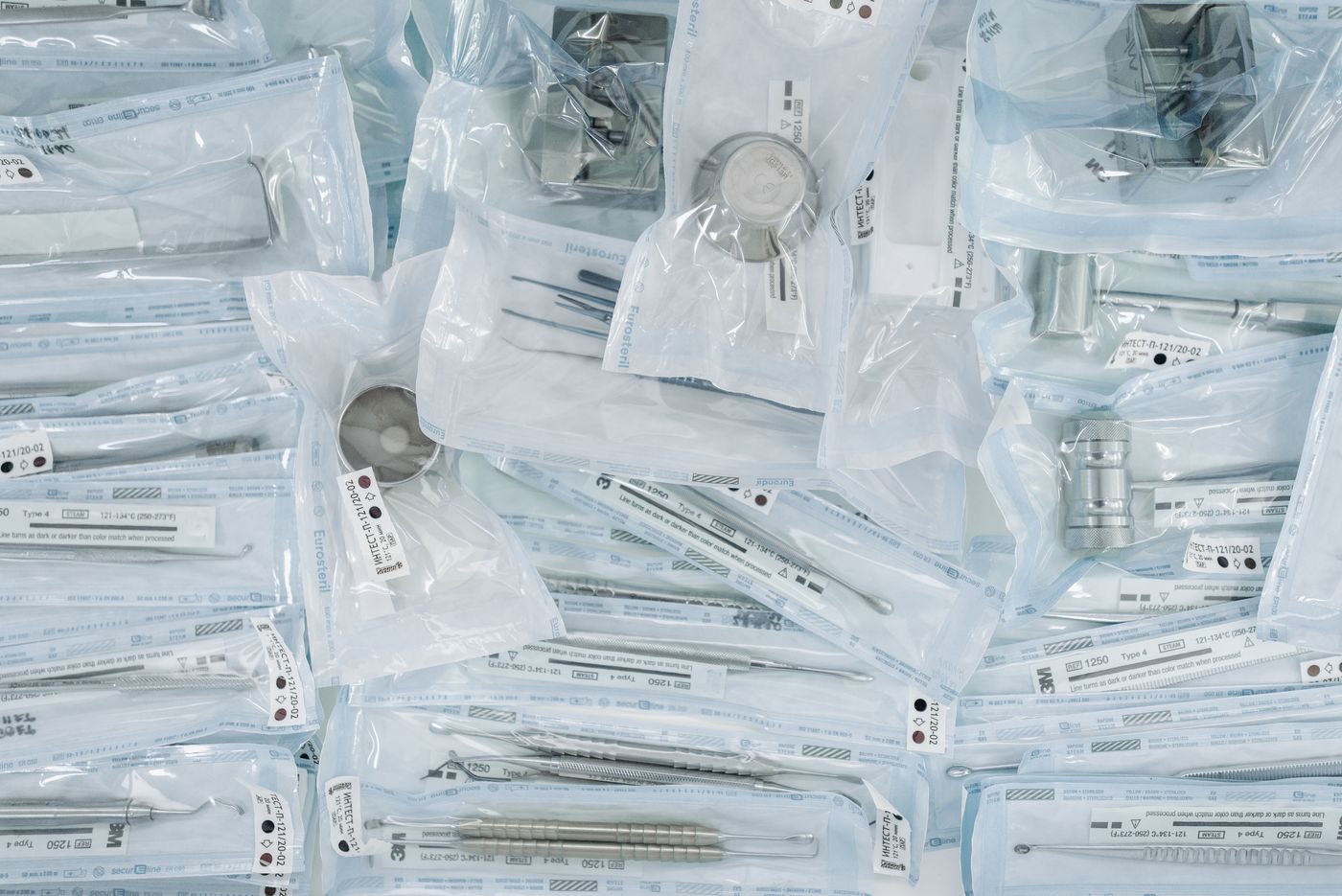Ein dichter Haufen sterilisierter Dental- und Chirurgieinstrumente, eingeschweißt in Sterilisationsbeutel aus Klarfolie und Papier, jeder Beutel bedruckt mit Referenznummern, Chargencodes und Dampfprozess-Indikatoren.