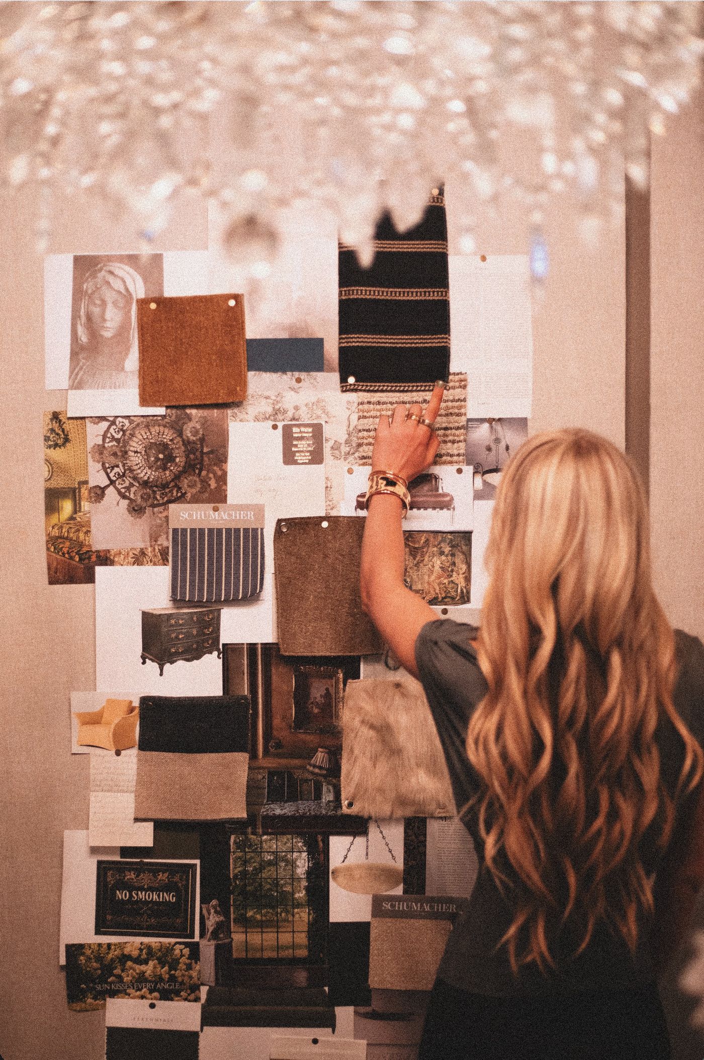 An interior designer reaches up to place a striped textile swatch on a large pinned board of furniture images, leather and fabric memos, and tearsheets.