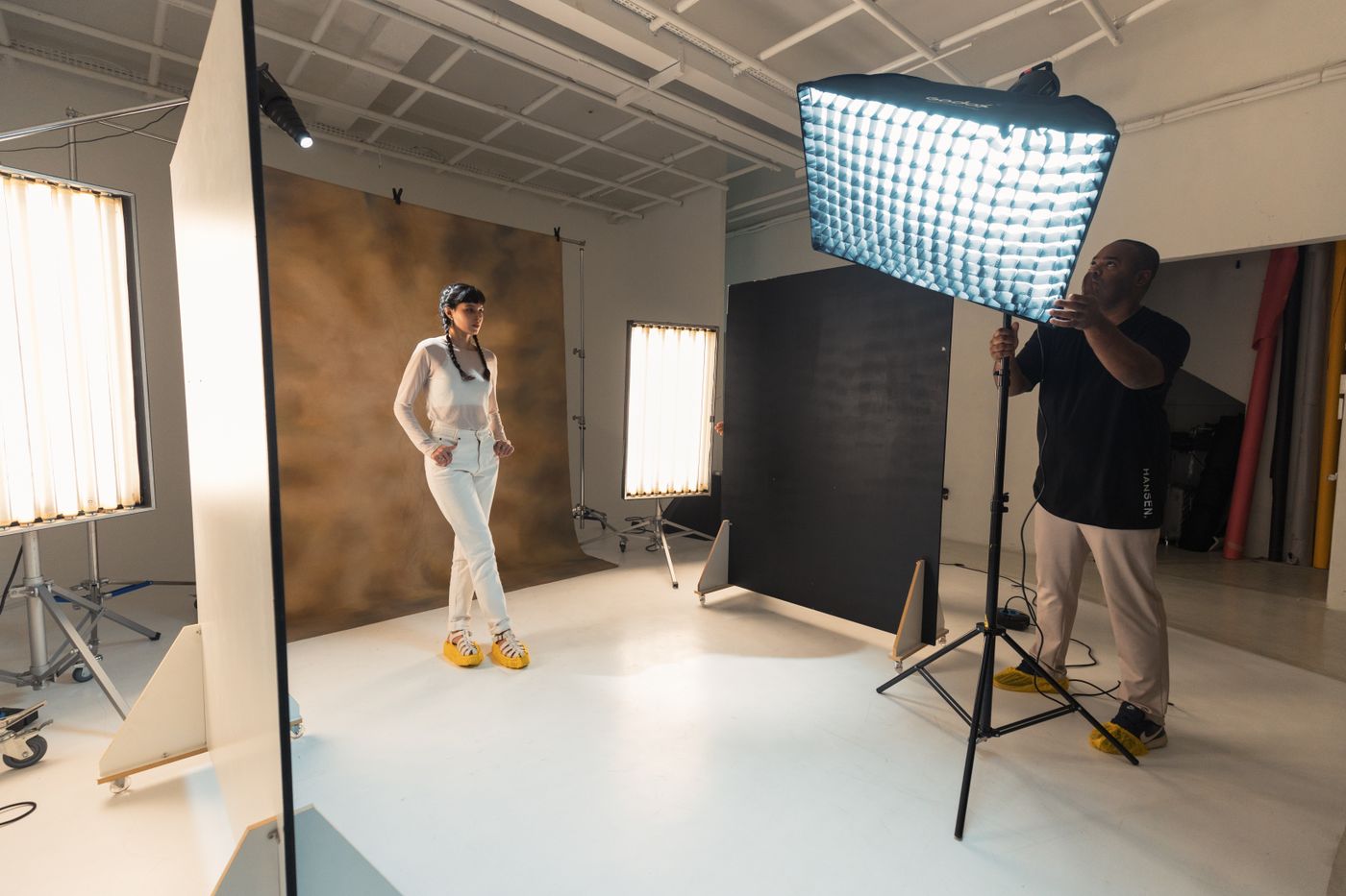 Un model stă la câțiva pași distanță de un fundal de studio suspendat, în timp ce un fotograf poziționează un panou softbox mare și dreptunghiular ca s-o lumineze uniform din față.