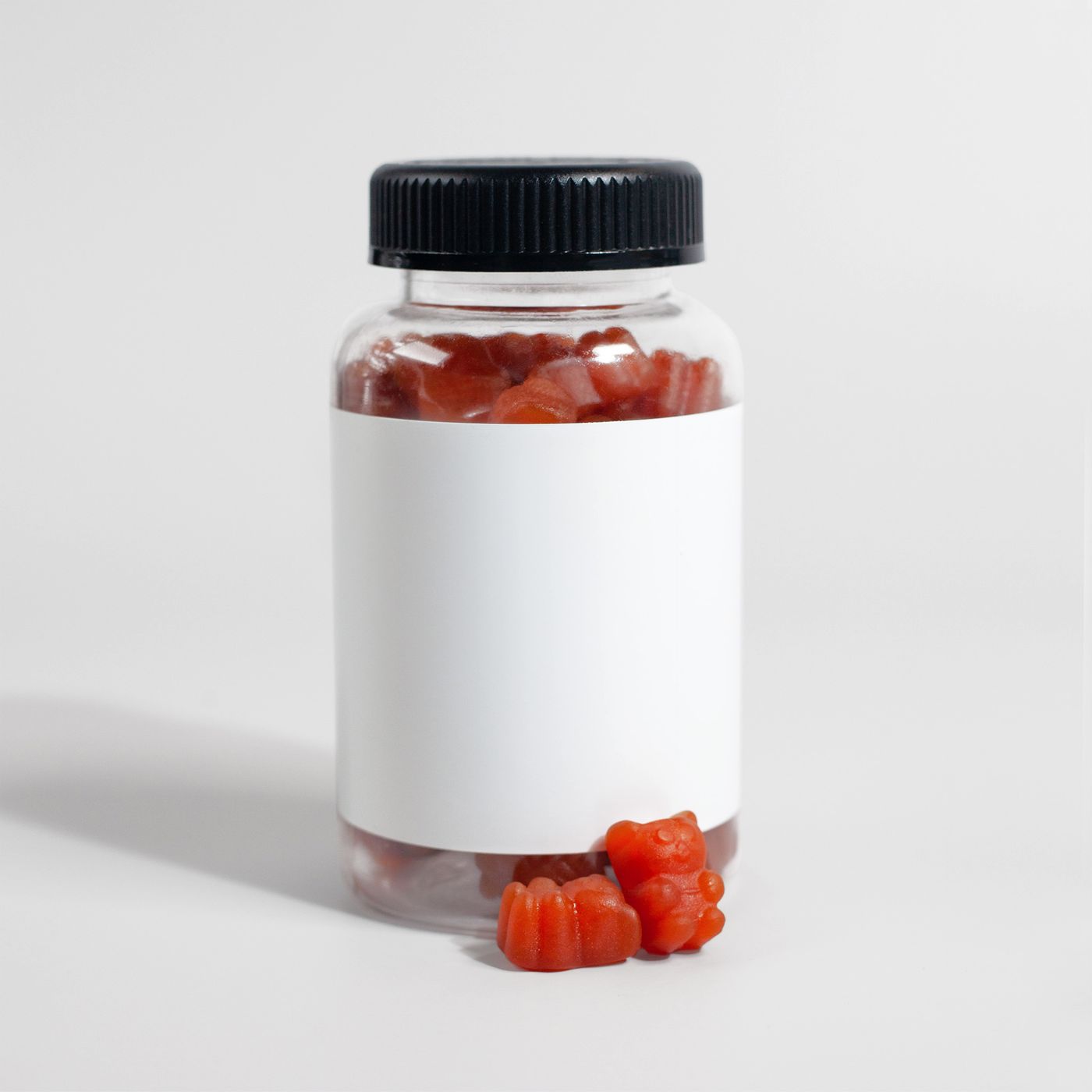 Un bote de plástico transparente con gominolas rojas, tapón de rosca negro y una etiqueta blanca sin imprimir, con tres gominolas sueltas al lado sobre una superficie blanca lisa.