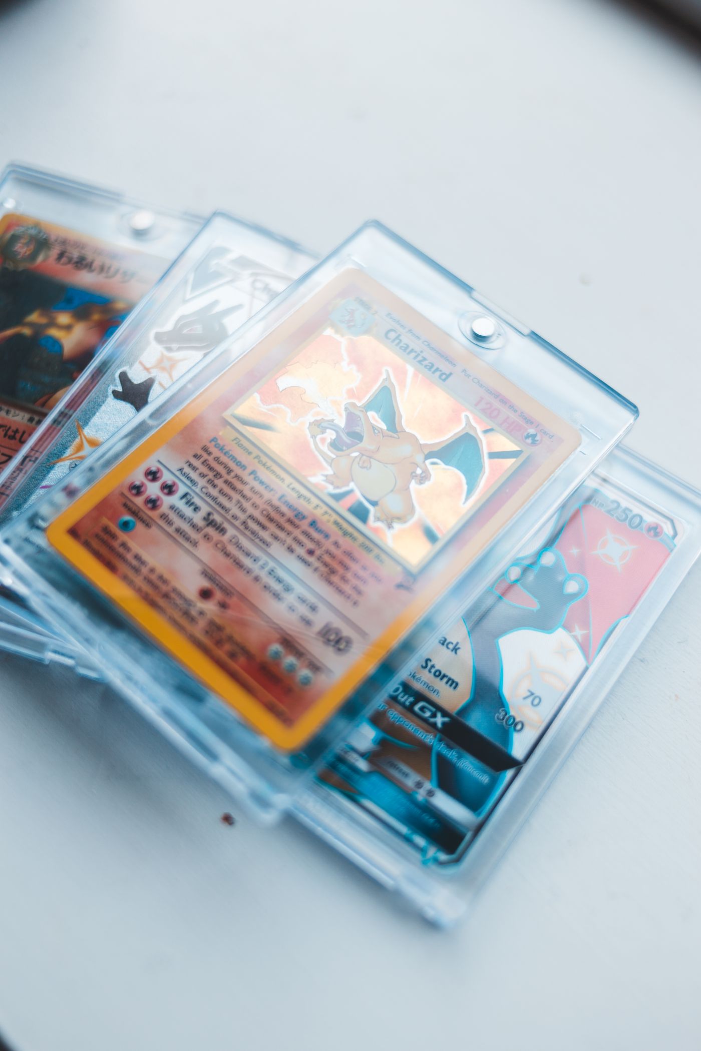 Kilka kart Pokemon, w tym Charizard, zamkniętych w przezroczystych, sztywnych plastikowych holderach na białym blacie, z mgiełką odbić i jasnymi refleksami na plastiku