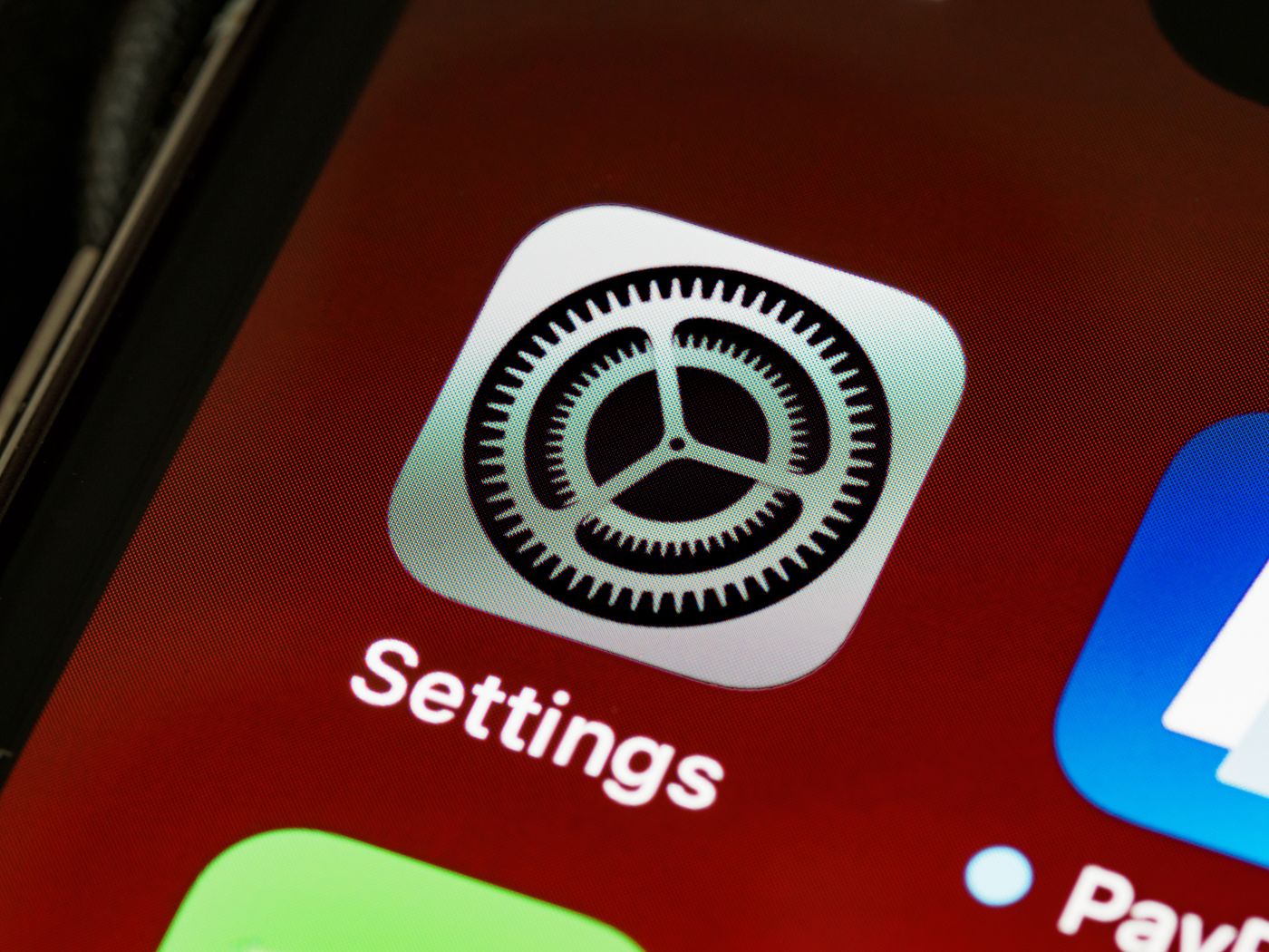 Extreme close-up of an iPhone home screen showing the grey Settings app icon with its cog wheel, labelled Settings, beside neighbouring app icons