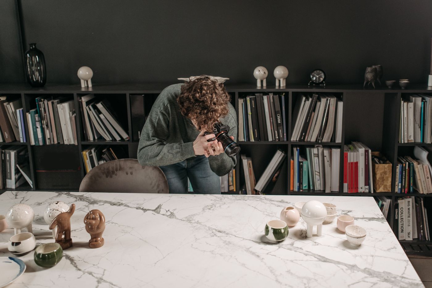 A photographer bends low over a large marble table, camera held close to a scattered group of small ceramic cups and figurines