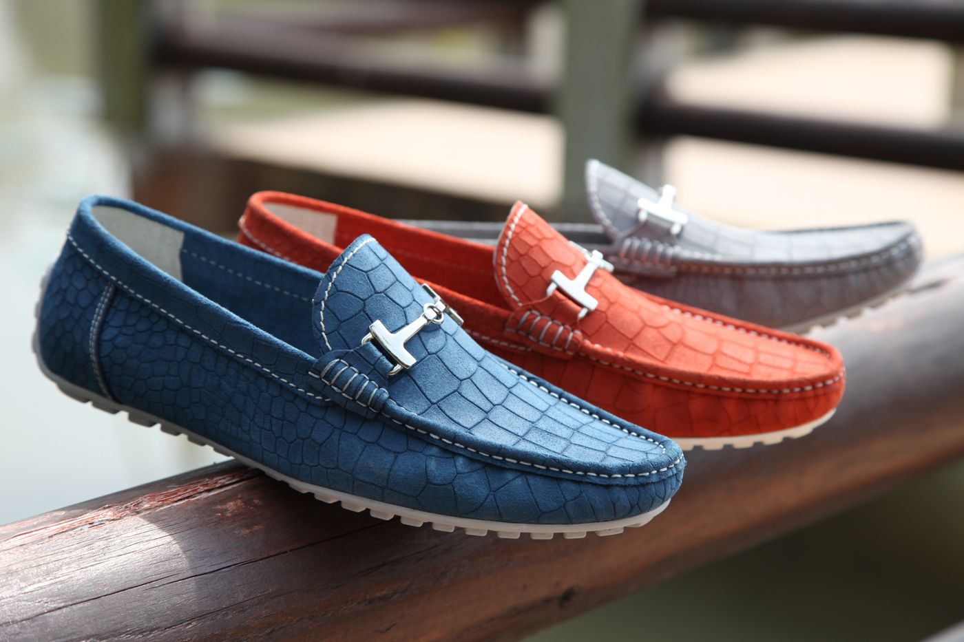 Three leather loafers in red, blue and grey lined up along a wooden railing, the nearest shoe sharply detailed while the ones behind it fall away into blur