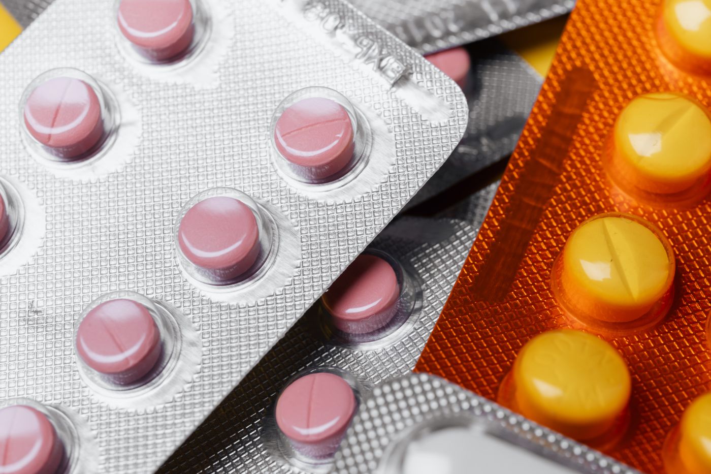 Nahaufnahme von Blisterpackungen: rosa Tabletten unter klaren Kuppeln auf geriffelter Silberfolie, daneben gelbe Tabletten in einer orangefarbenen Folienpackung, mit harten hellen Reflexen quer über Kuppeln und Rillen.