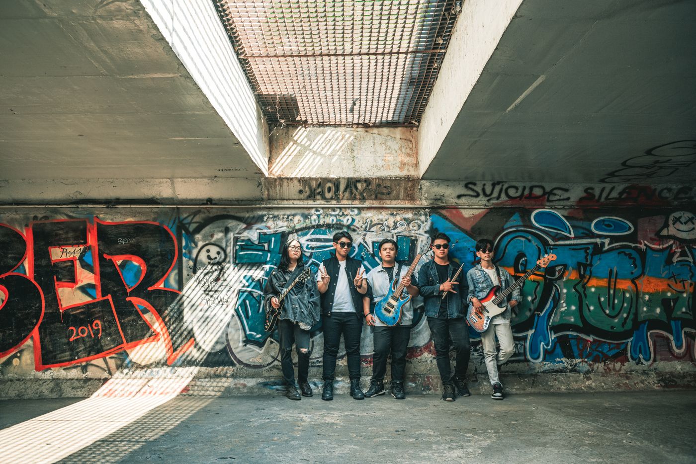 Alt geçit altında grafitili beton duvar önünde omuz omuza dizilmiş gitarlı beş müzisyen, tek yandan yumuşak gün ışığı