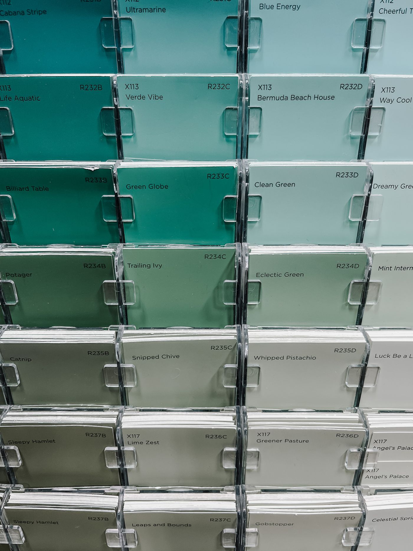 A close-up of colour swatch cards laid out in rows of green, sage and pale grey-green, each card carrying a printed reference code beside its marketing name.