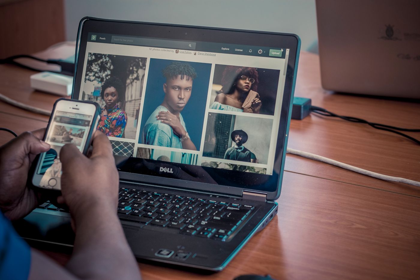 Hands holding a smartphone showing photographs, held up in front of an open laptop whose screen displays the same kind of photo gallery in a browser