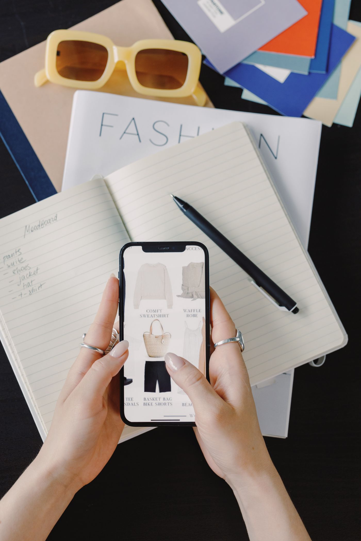 Dwie dłonie trzymają smartfon z siatką produktów w aplikacji zakupowej — małe kafelki miniatur z bluzą, szlafrokiem, słomianą torbą i szortami pod krótkimi podpisami