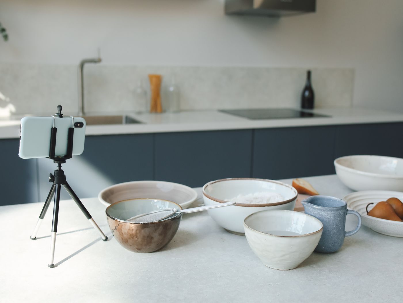 A smartphone mounted on a small tripod aimed at ceramic bowls and a mug arranged on a bright kitchen counter