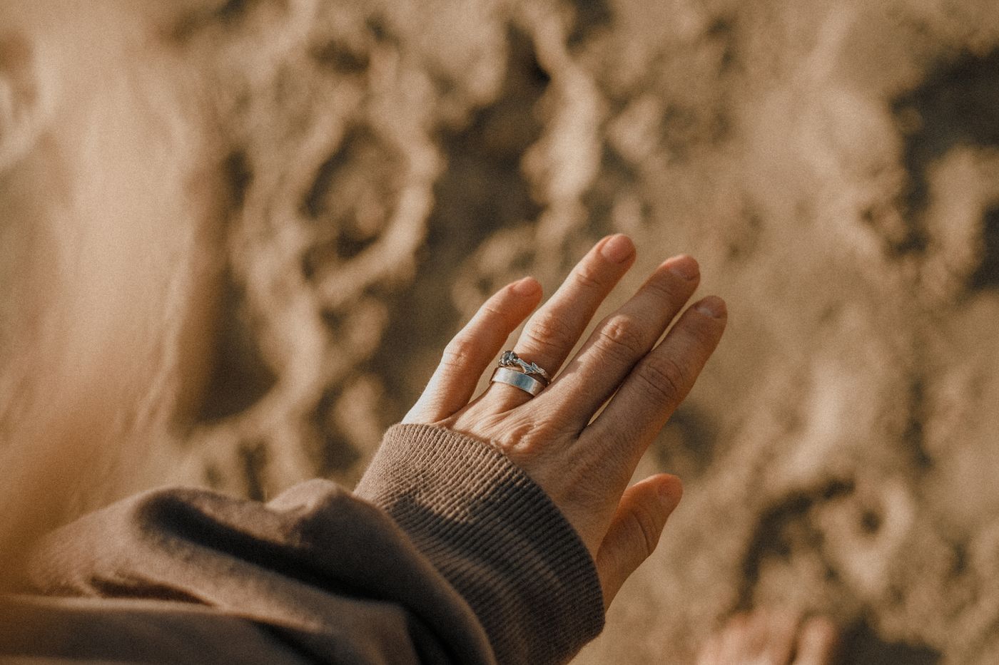 A woman's hand resting above sand, wearing a slim engagement ring and wedding band, photographed casually from above