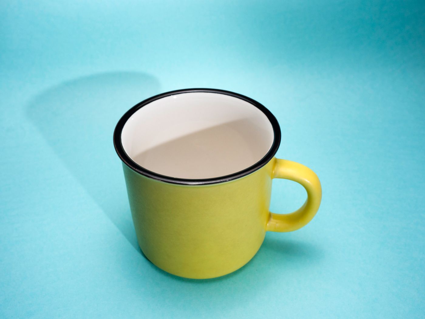 A single empty yellow mug with a black rim and white interior, shot from a raised angle on a plain light blue background with bare space and a soft shadow around it.