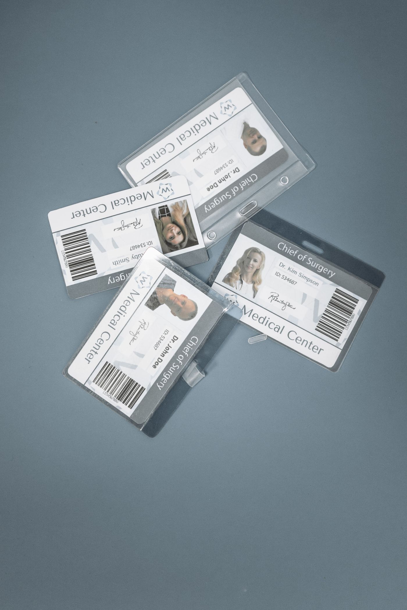 Patru legitimații de serviciu plastifiate, așezate pe un birou gri, fiecare tipărită cu o poză mică de portret, un nume, un număr de identificare și un cod de bare.