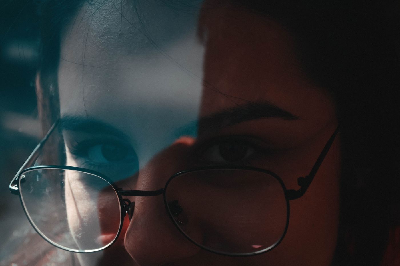 Extreme close-up of a woman's face in thin metal-framed glasses; her eye is clearly visible through the lens on one side while a bright reflection of the scene in front of her spreads across the other side of the frame