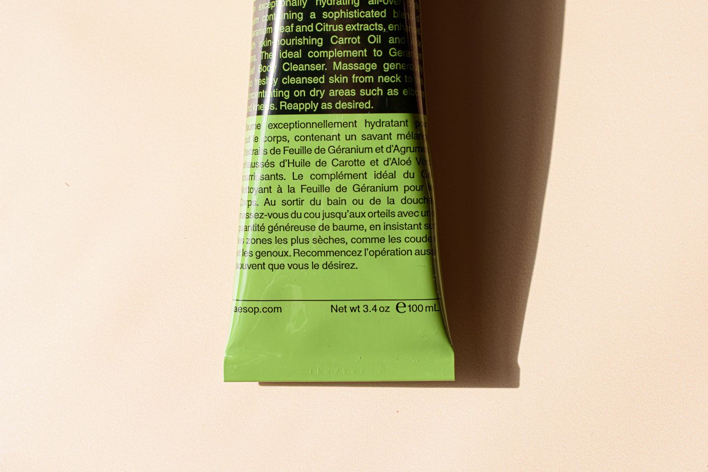 Primer plano del reverso de un tubo cosmético verde con instrucciones de uso impresas en inglés y francés, encima de una dirección web y un peso neto de 100 mL.