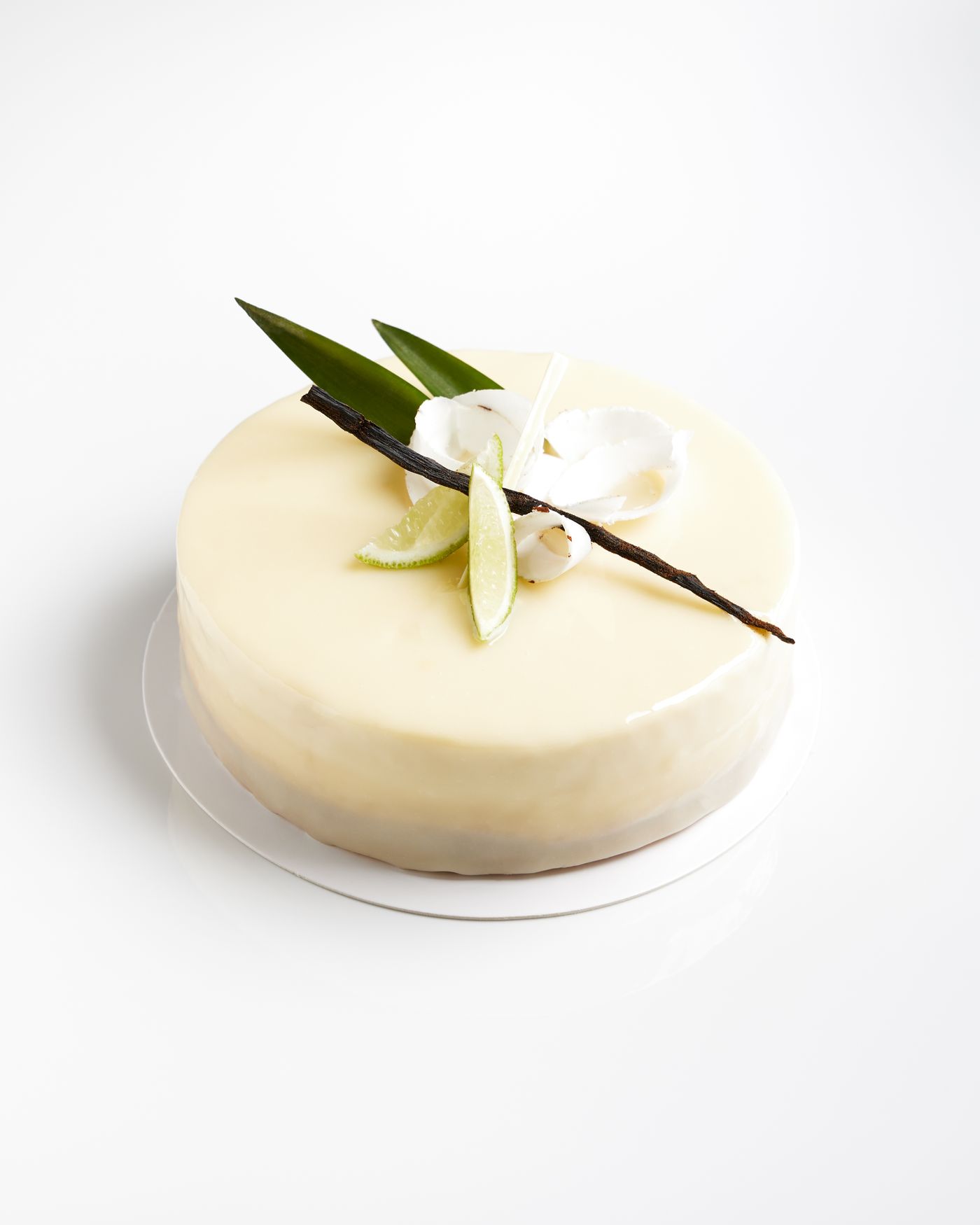 A round cake with glossy pale glaze, topped with a vanilla pod, lime wedges and coconut curls, sits on a white board against a seamless white background with a soft shadow gathered tightly beneath its base.