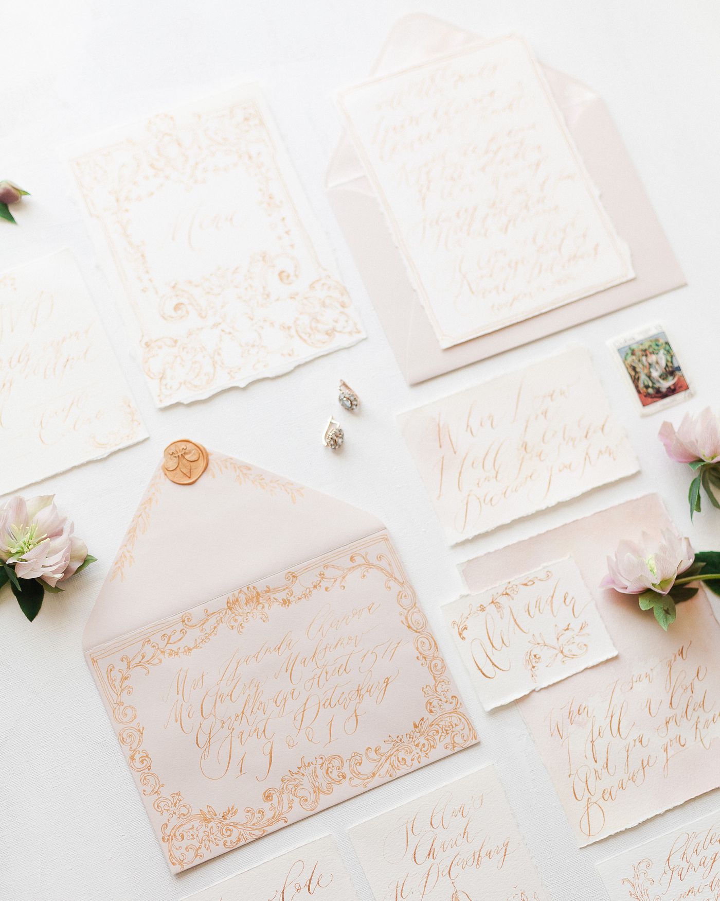 An overhead flat lay of blush wedding stationery: a wax-sealed envelope with gold calligraphy, deckle-edged invitation cards, a postage stamp, earrings and hellebore blooms arranged on a plain white surface.