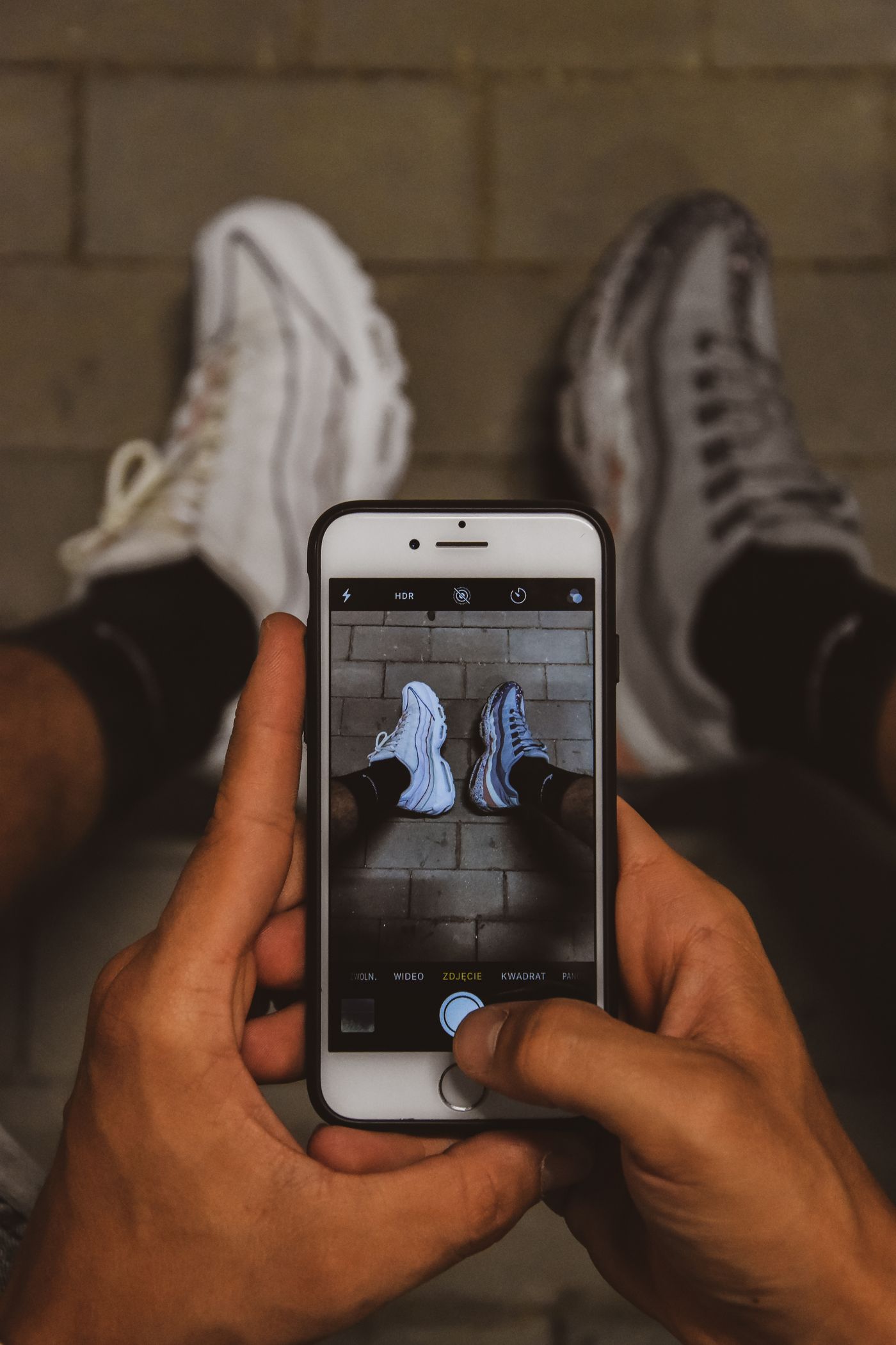 A person photographing their white sneakers with a smartphone, shooting down at the shoes on brick pavement
