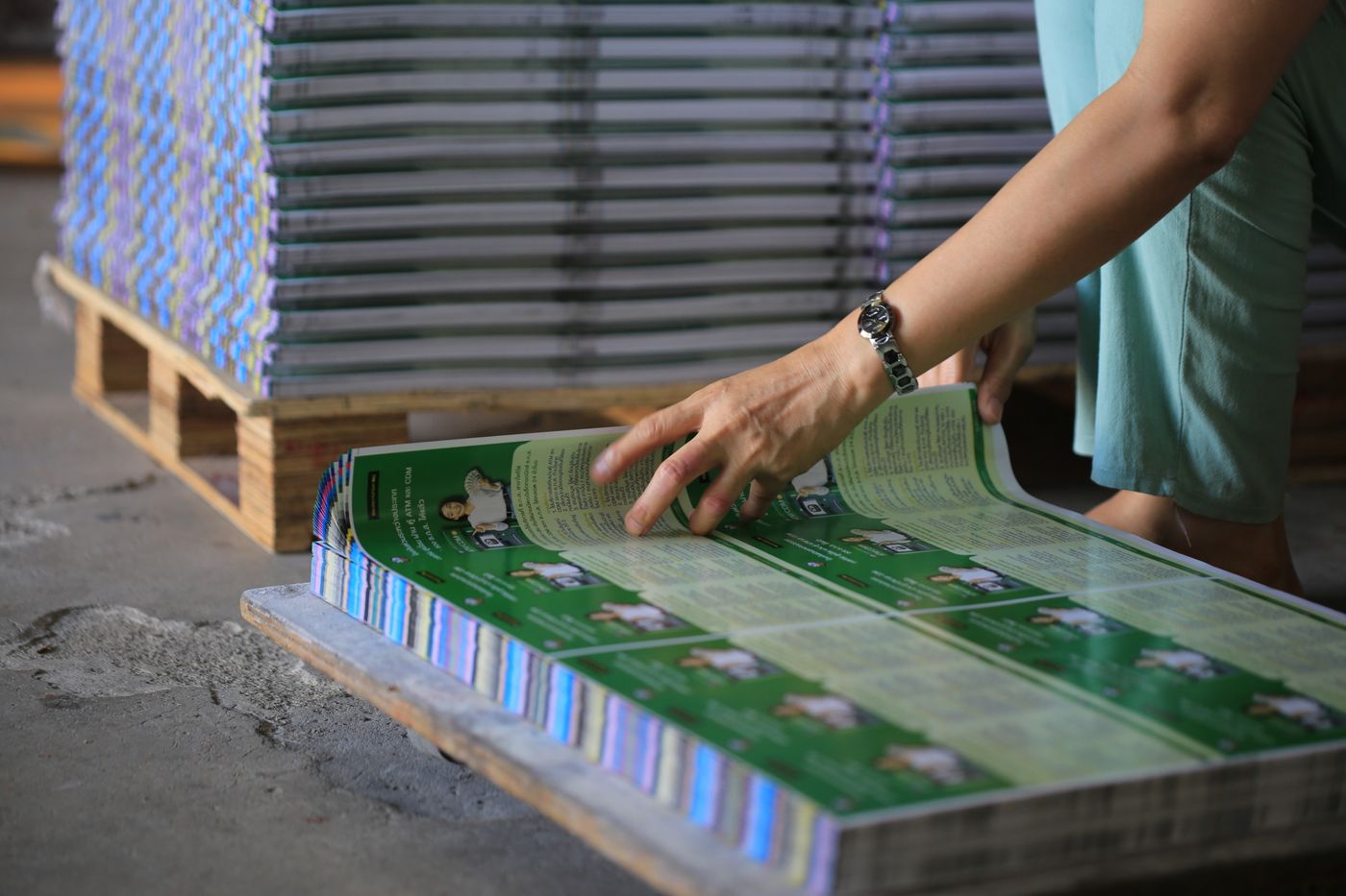 A hand turning the pages of a freshly printed booklet on a pallet stacked with finished copies in a print shop