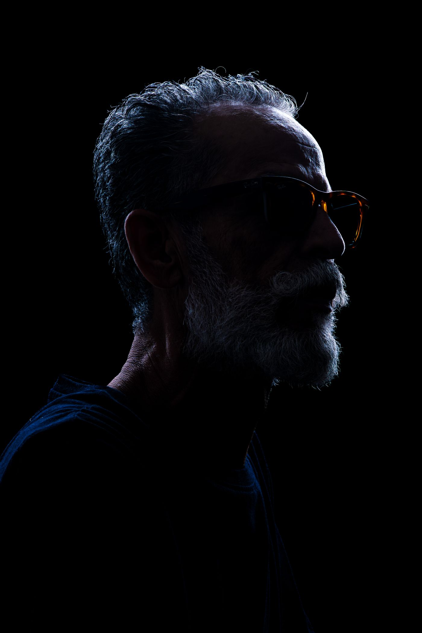 A bearded man seen in profile against a black background, with a bright pale rim of light tracing the outline of his hair, beard and shoulder.