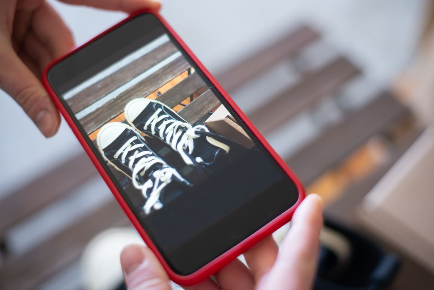 Close-up of a person's hands holding a smartphone, the screen filled with a just-taken photo of a pair of black sneakers on a bench