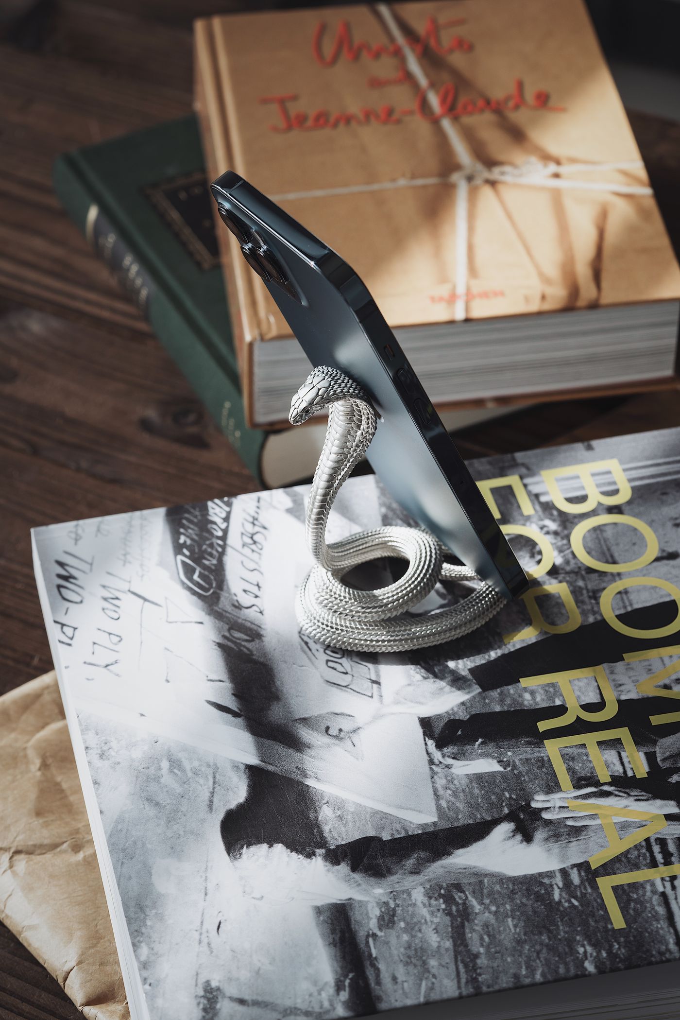 Un telefono in verticale dentro un supporto di metallo che poggia su una pila di libri rilegati e volumi fotografici, su un tavolo di legno, senza mani a sostenerlo.