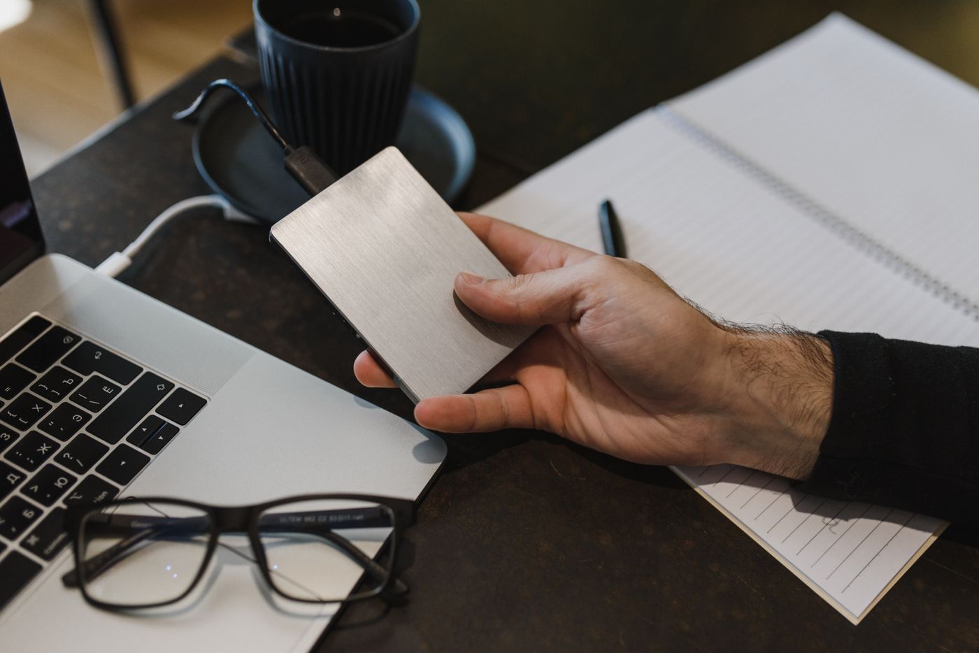 A man holds a portable external hard drive plugged into a laptop on a desk, a coffee cup and glasses beside it