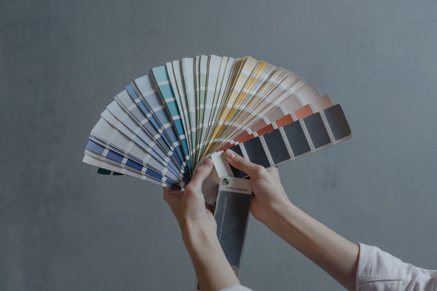 Two hands holding a fanned-out deck of paint colour swatch cards in blues, greens, yellows and greys against a grey wall