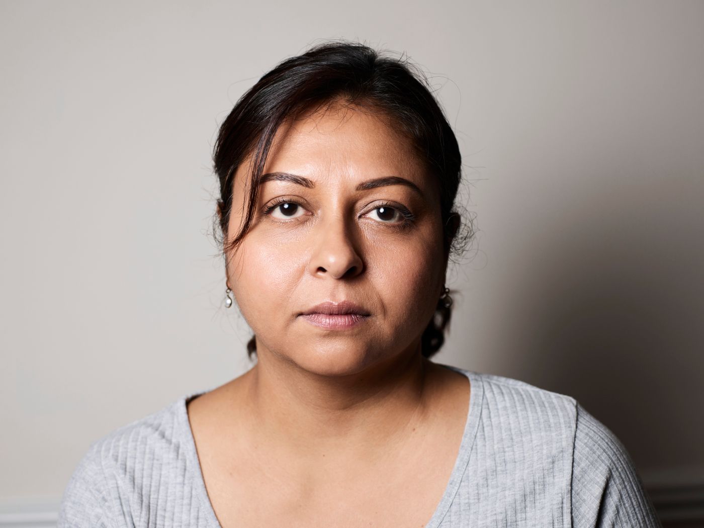 Head-and-shoulders portrait of a woman in a grey top looking straight at the camera against a plain, evenly lit grey wall