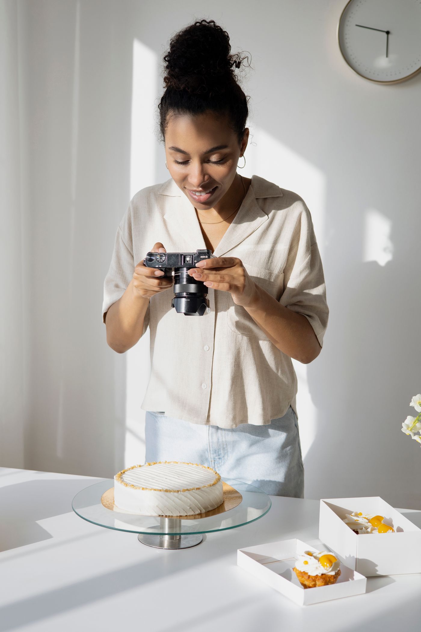 A woman in a linen shirt leans over a white worktop and photographs a round white-iced cake on a glass stand with a small mirrorless camera, in bright directional daylight from a nearby window.