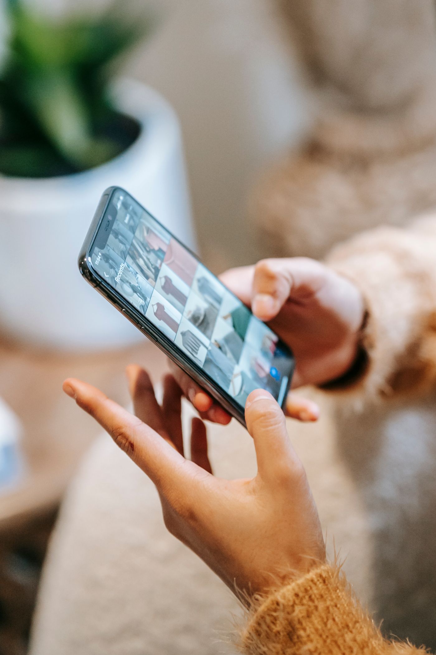 A woman's hand holding a smartphone that shows a grid of photos in the gallery app, with a blurred living room behind it