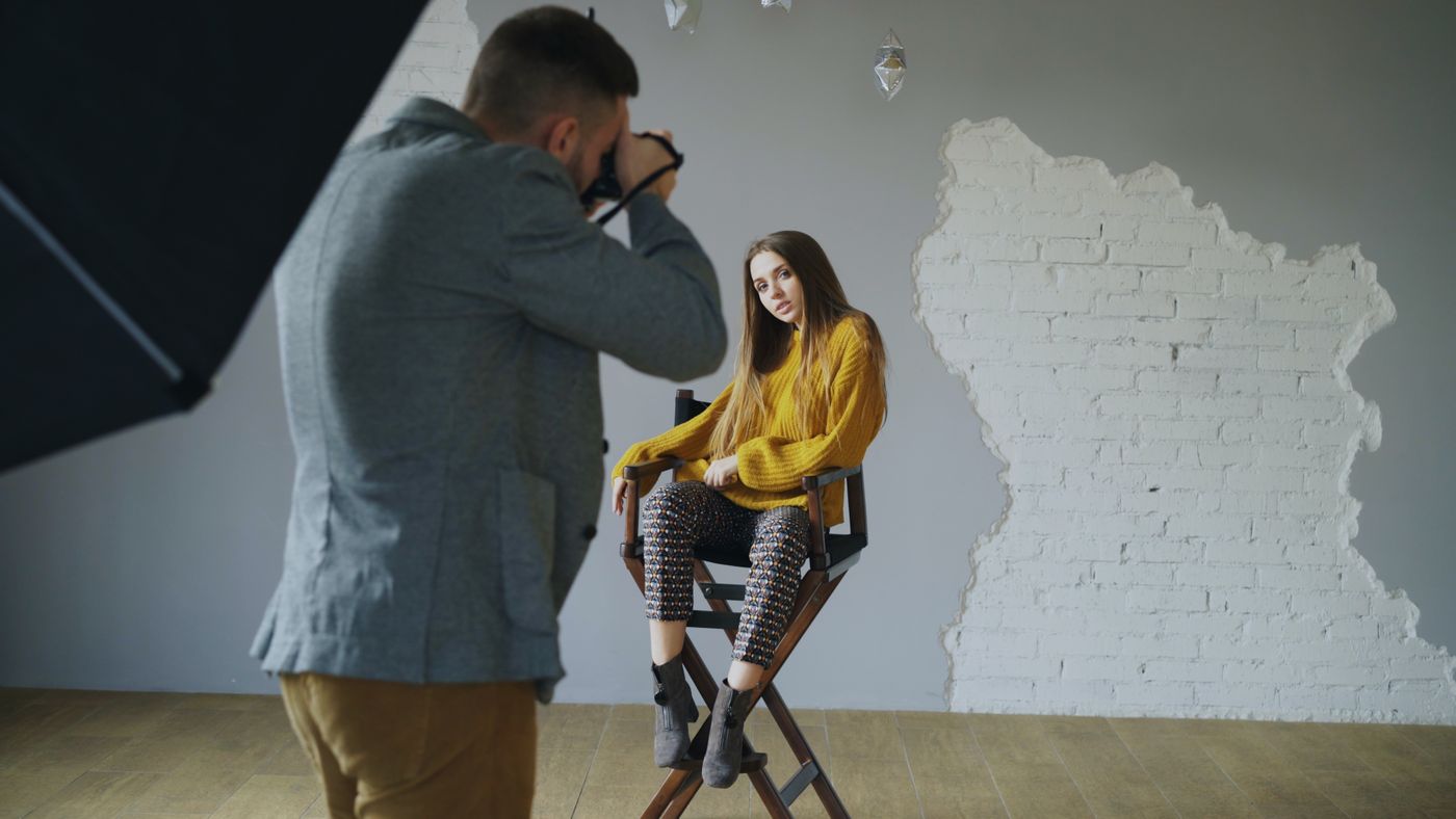 Un fotograf cu aparatul la ochi fotografiază un subiect așezat, luminat de un softbox mare, pe un perete alb gol