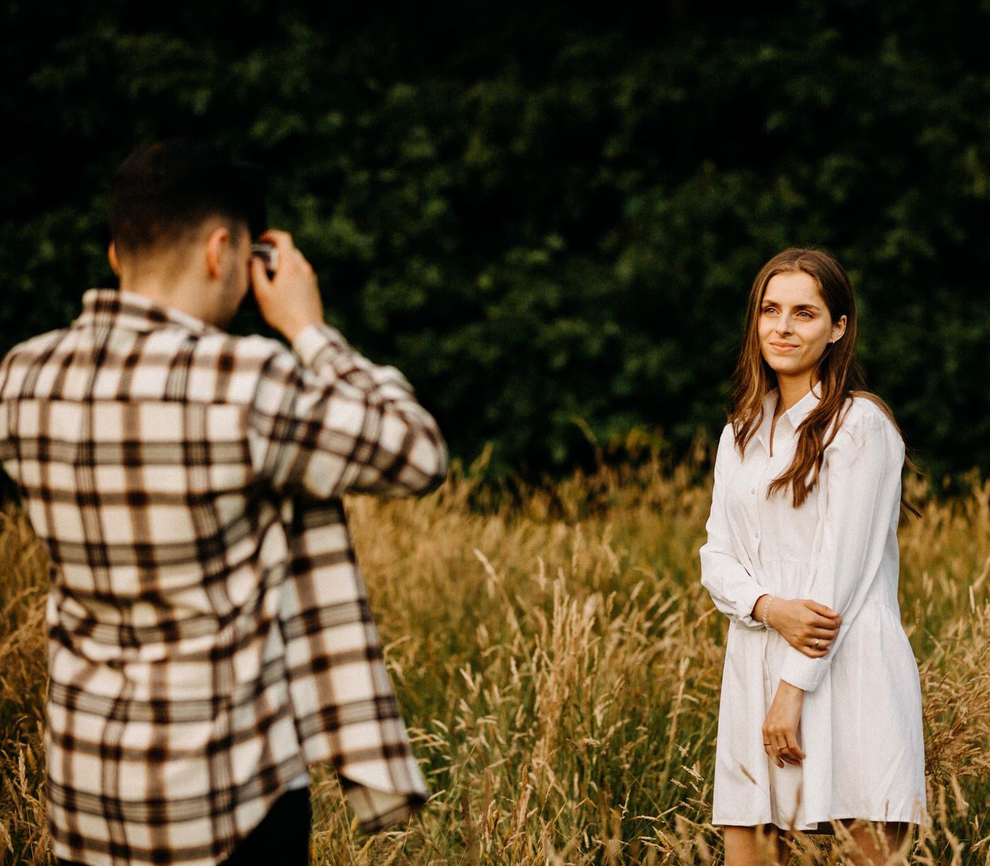 A man photographing a smiling woman who stands several metres away in a grassy field, with the dark treeline behind her falling out of focus.