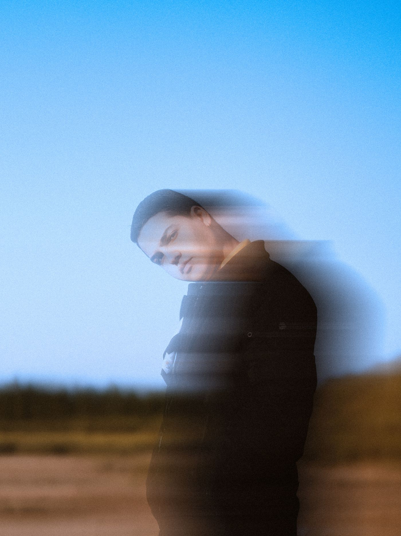 A person standing outdoors against a clear sky, photographed with a slow shutter so their head and shoulders smear into a soft, doubled blur with no defined outline.