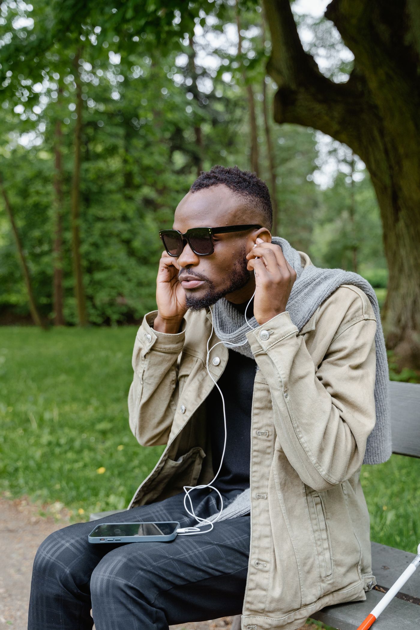 A man in a beige jacket and dark sunglasses sits on a park bench fitting white wired earbuds into his ears, his smartphone lying on his lap and a white cane resting beside him.