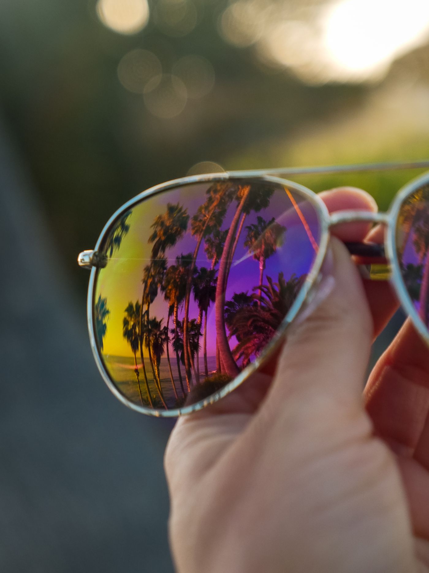 A hand holding up mirrored aviator sunglasses whose lens is completely filled with a reflected image of tall palm trees along a coastline at sunset