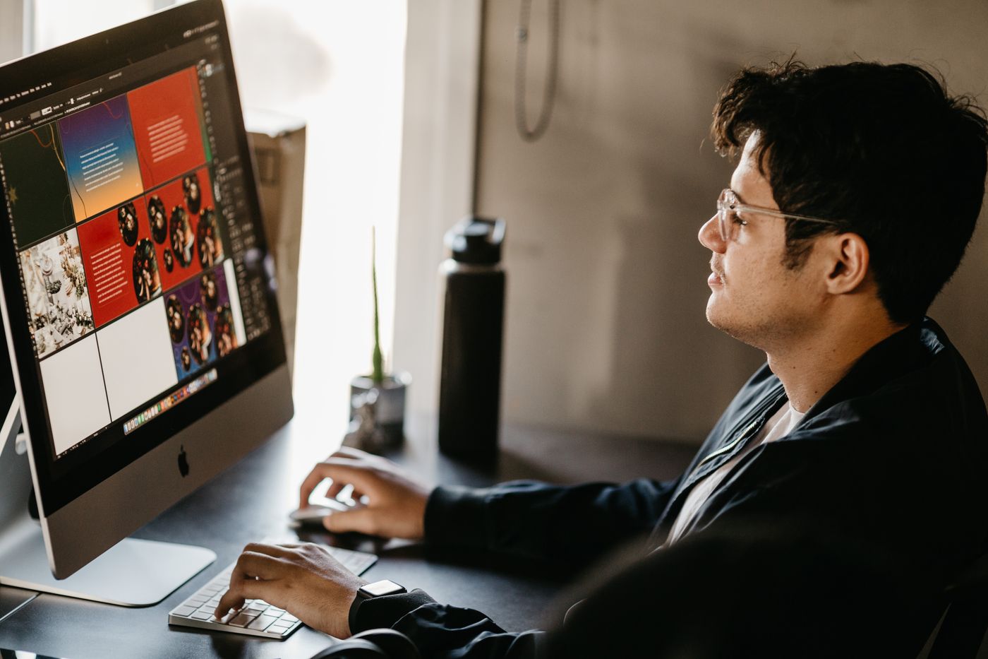 A graphic designer in glasses sitting at a desktop computer in a modern office, working on visual artwork displayed on the monitor