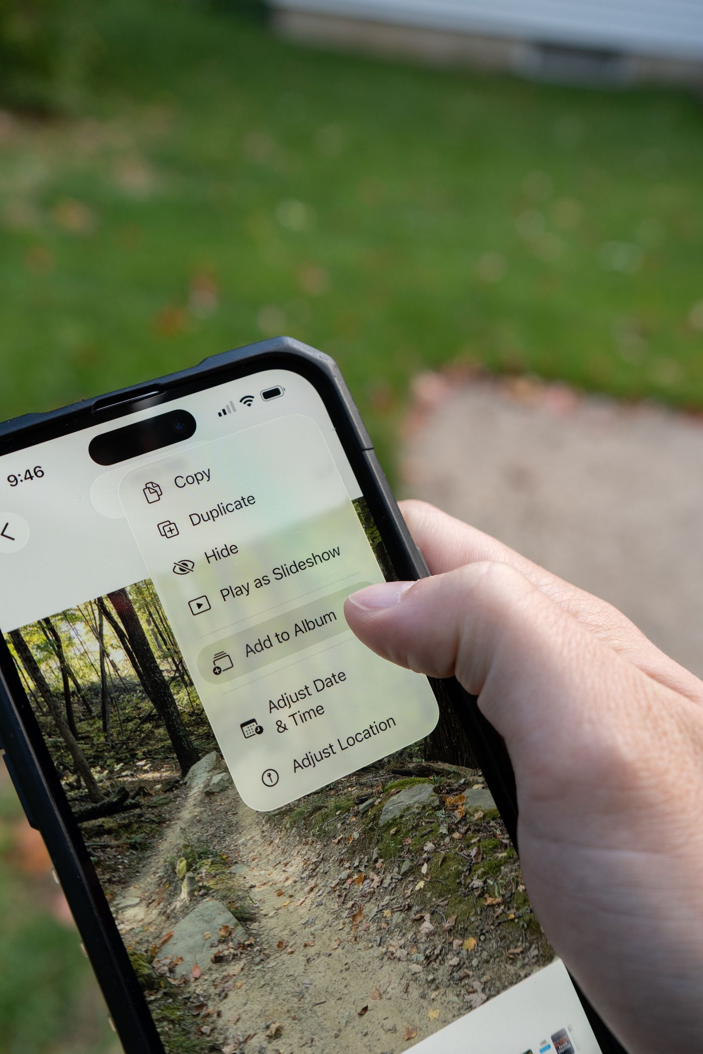 A thumb pressing an iPhone screen where a long-press context menu is open over a photo, listing options such as Copy, Duplicate, Hide and Add to Album