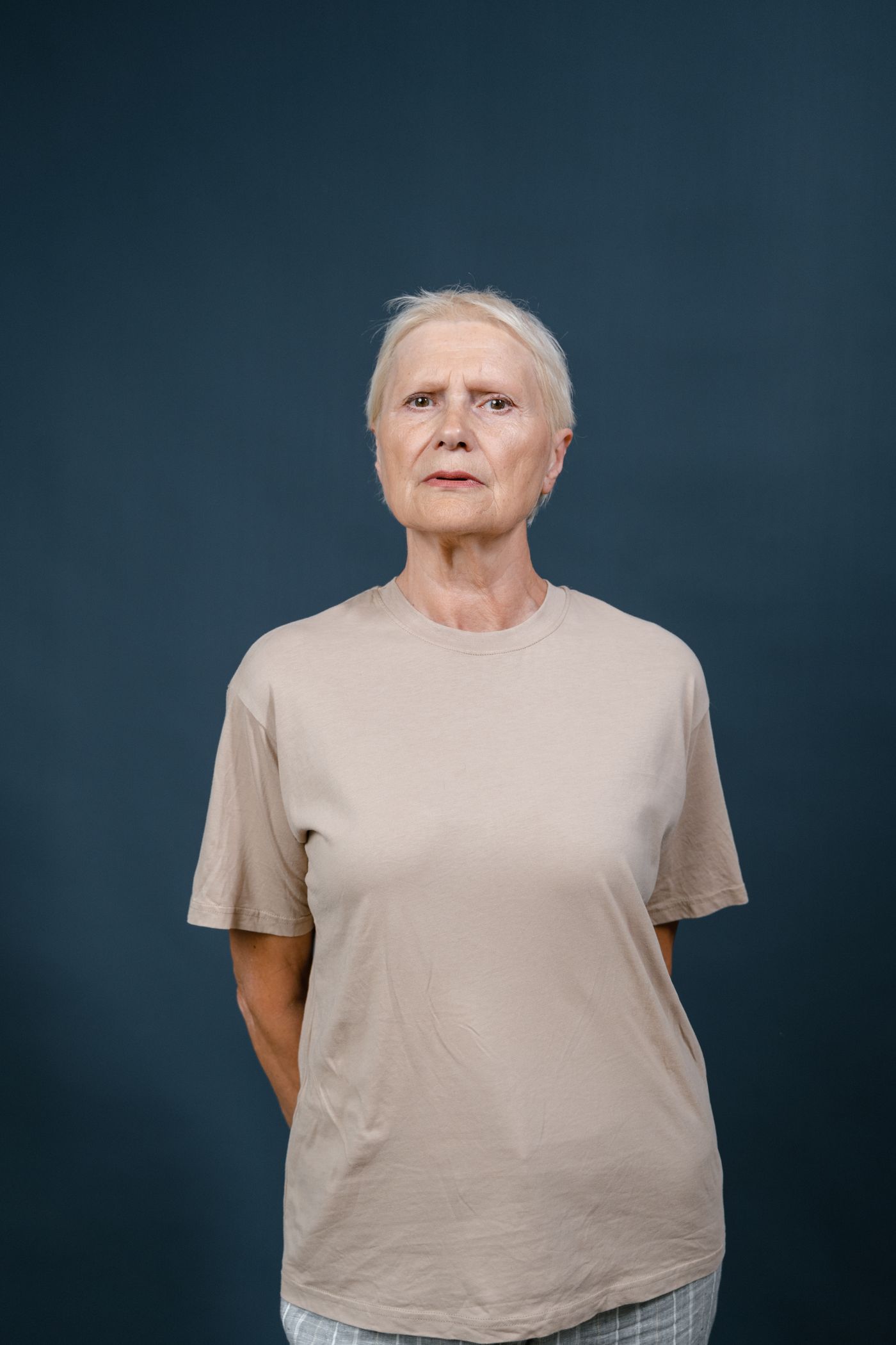 An elderly woman with short silver hair, photographed from the chest up against a flat dark blue-grey studio backdrop.