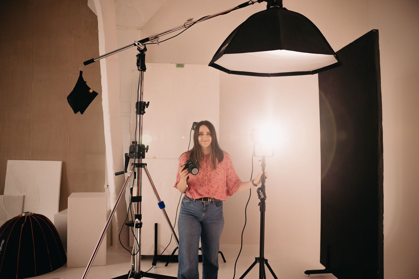 A photographer holding a camera in a brightly lit studio, standing beside a large softbox on a stand that is angled in from one side of the shooting position