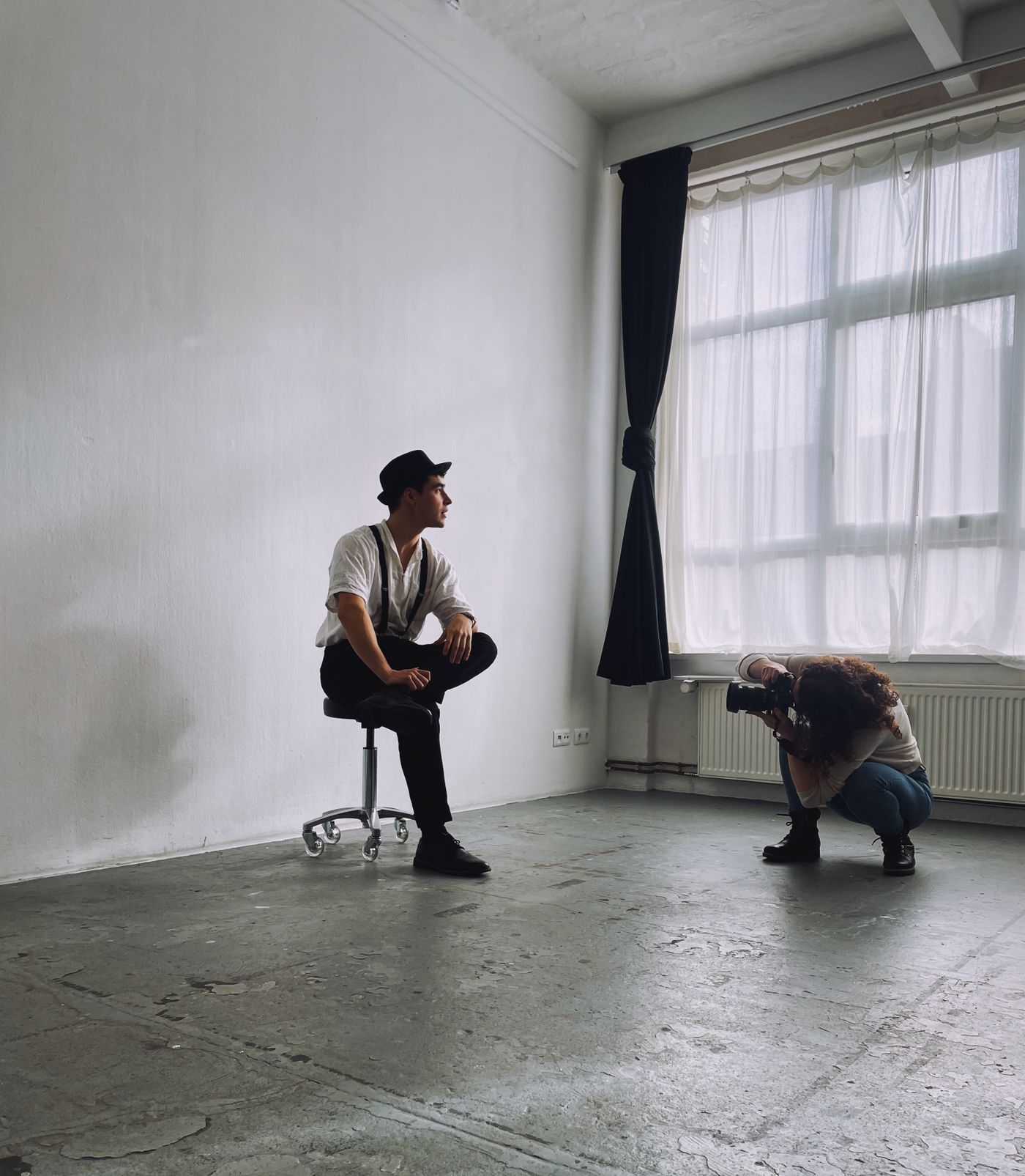 A photographer crouching by a curtained window photographs a man seated on a stool against a bare white wall in an empty room