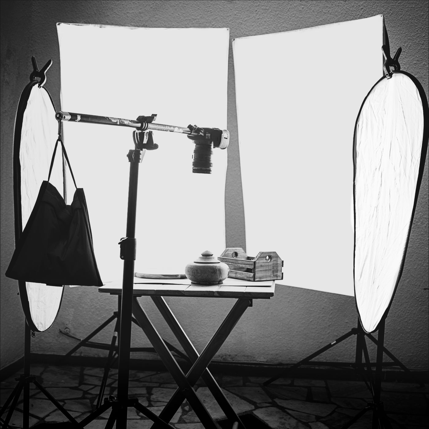 A small studio rig with a camera on a boom arm pointing down at a lidded pot and a wooden box on a folding table, flanked by diffusion panels and reflectors
