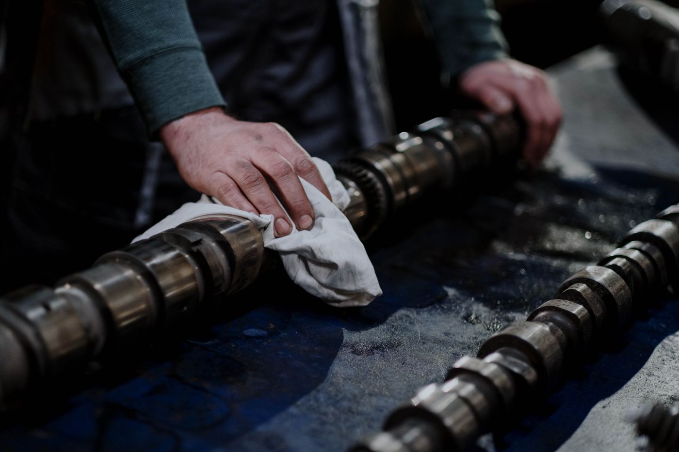 Hands wiping a steel camshaft with a white cloth on a dark oil-stained workbench, with two more camshafts alongside