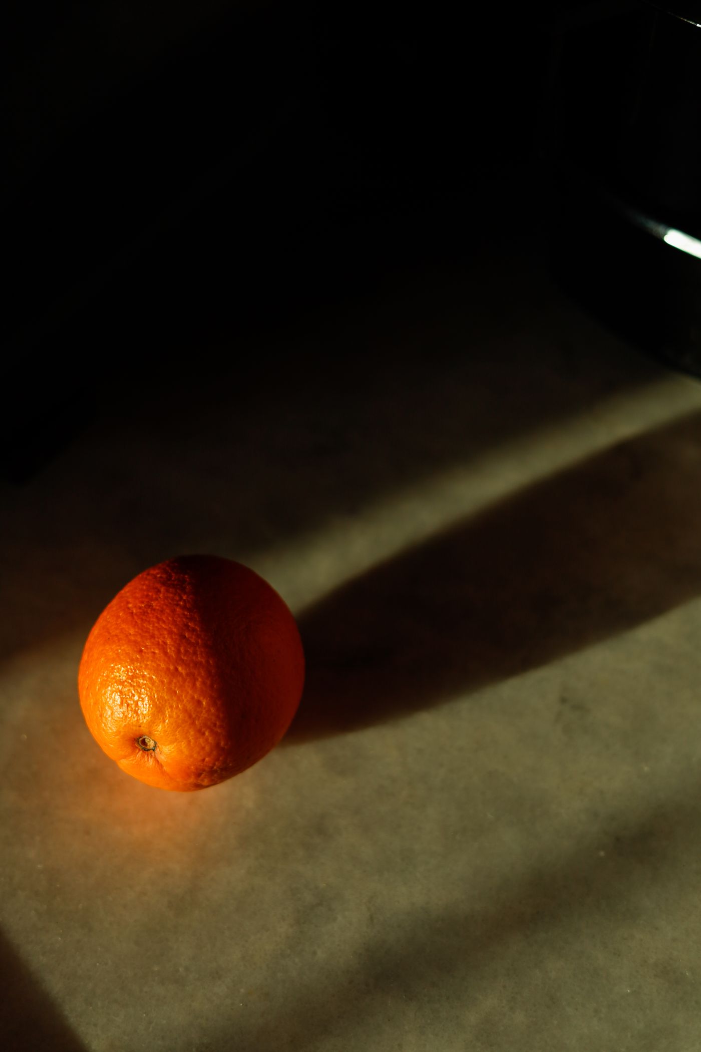 O singură portocală stă pe o masă în lumină dură de soare, cu umbra întinzându-se pe suprafață alături de ea, nu peste fruct.