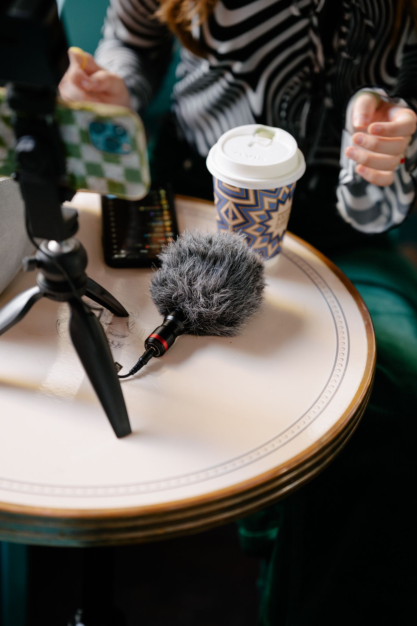 A smartphone mounted on a tripod stands on a table with its lens facing a patterned coffee cup, a microphone set up alongside it.
